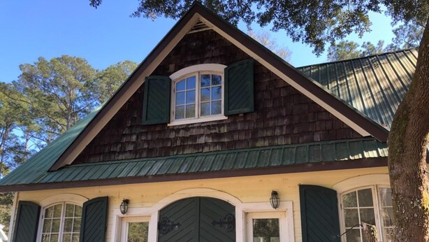 Une maison traditionnelle en bois, dotée d’un toit à pignon, d’une entrée centrale avec une grande porte et de volets verts aux fenêtres.