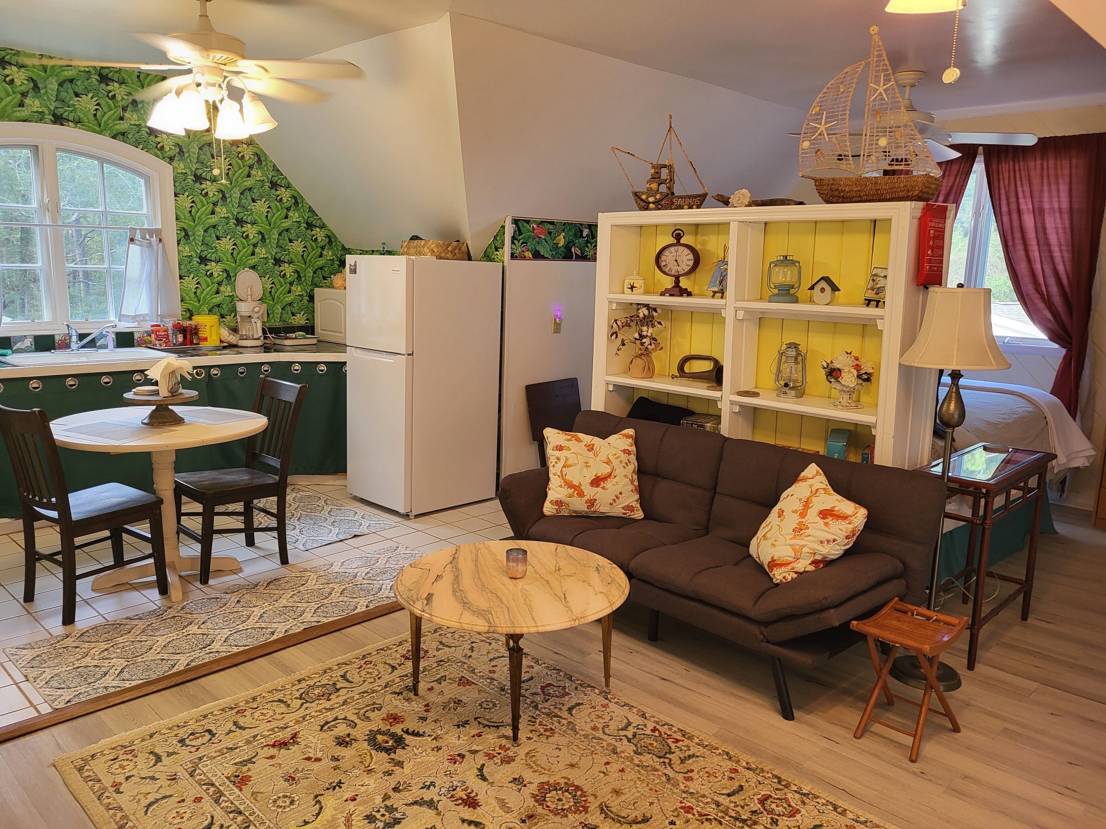 Una sala de estar acogedora con un sofá marrón, una mesita redonda y una zona de cocina con refrigerador y ventana.