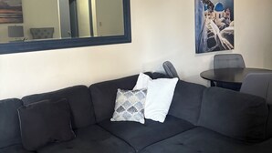 Un soggiorno moderno con un divano modulare nero, un tavolino in vetro, uno specchio decorativo e un quadro incorniciato raffigurante una scena costiera.