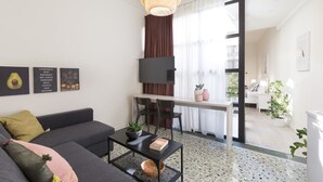 En moderne stue med en sofa, et sofabord og et fjernsyn på et stativ. Der er et spiseområde med et bord og stole, og et køkken er synligt i baggrunden.
