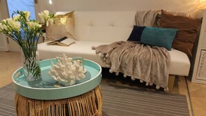 Una sala de estar con un sofá blanco, una mesa de centro redonda de mimbre con una bandeja azul turquesa, un jarrón con flores blancas y una pieza decorativa de coral.