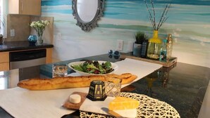 Una cocina con encimera de mármol, un cuadro del océano, un espejo decorativo y un jarrón con ramas.