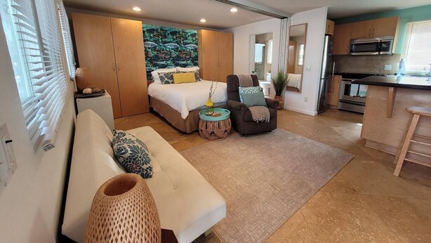 Una habitación compacta y moderna con cama, sofá, una mesita redonda y una zona de cocina con microondas y horno.