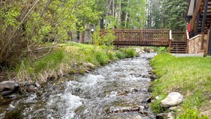 Pieni, kirkasvetinen puro virtaa luonnonkauniissa ympäristössä, ja sen yllä on puinen silta sekä sen rannoilla kivimuurit.