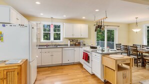 A modern kitchen with white cabinets, a wooden dining table, and a view of the outdoors. - River View House (Center Conway)