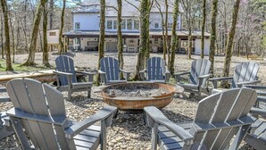 A rustic outdoor seating area with Adirondack chairs arranged around a fire pit, set against a backdrop of trees and a house. - Chasing Rainbows Lodge - 6 King Suites (Broken Bow)