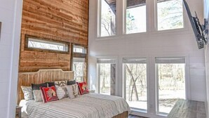 A bedroom with a wooden headboard, a bed with patterned bedding, and multiple windows providing natural light. - Chasing Rainbows Lodge - 6 King Suites (Broken Bow)