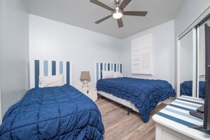 A bedroom with two beds, a ceiling fan, a bedside table with a lamp, and a window with blinds.