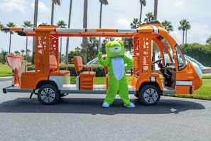 A mascot in a green frog costume standing next to a vehicle with a ladder and orange seats.