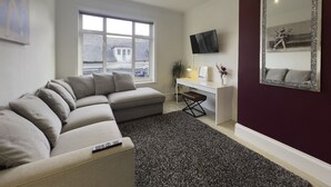 A modern living room with a sectional sofa, a desk, a large mirror, and a TV on the wall.