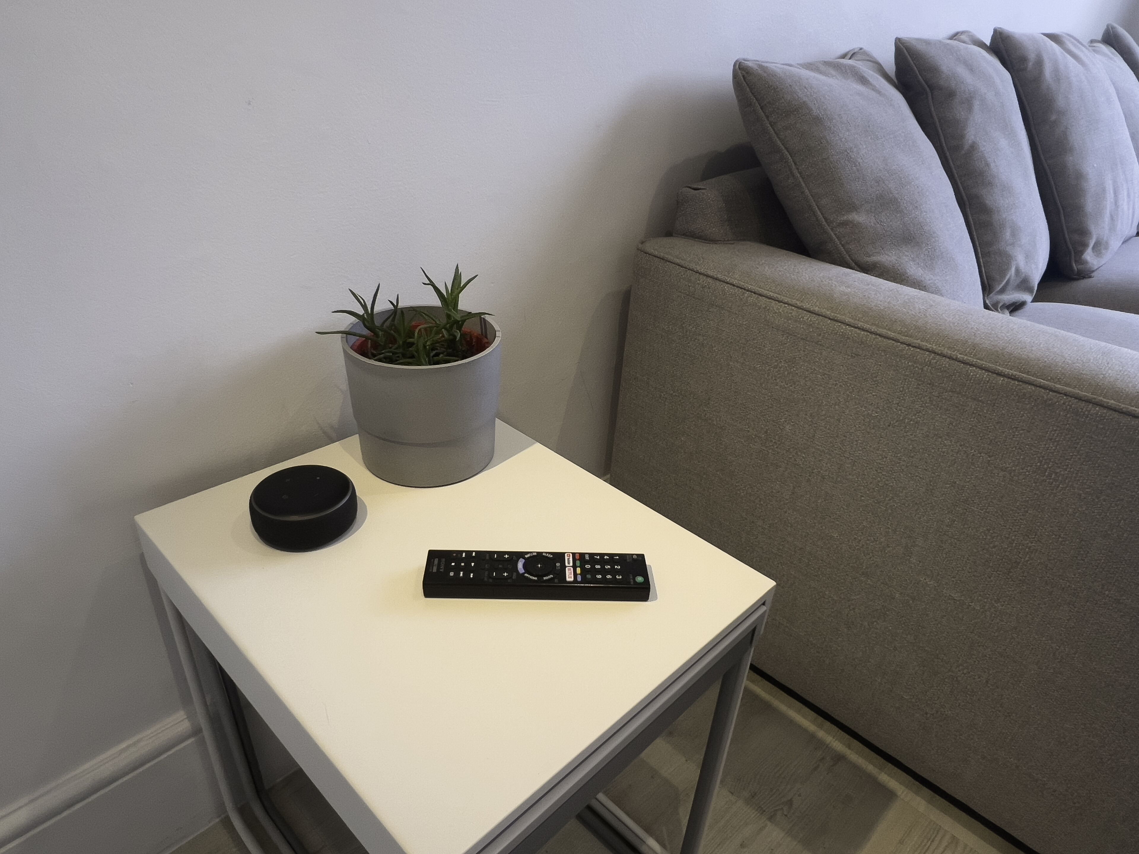 Una taula auxiliar petita de color blanc amb un comandament a distància i una planta en test.
