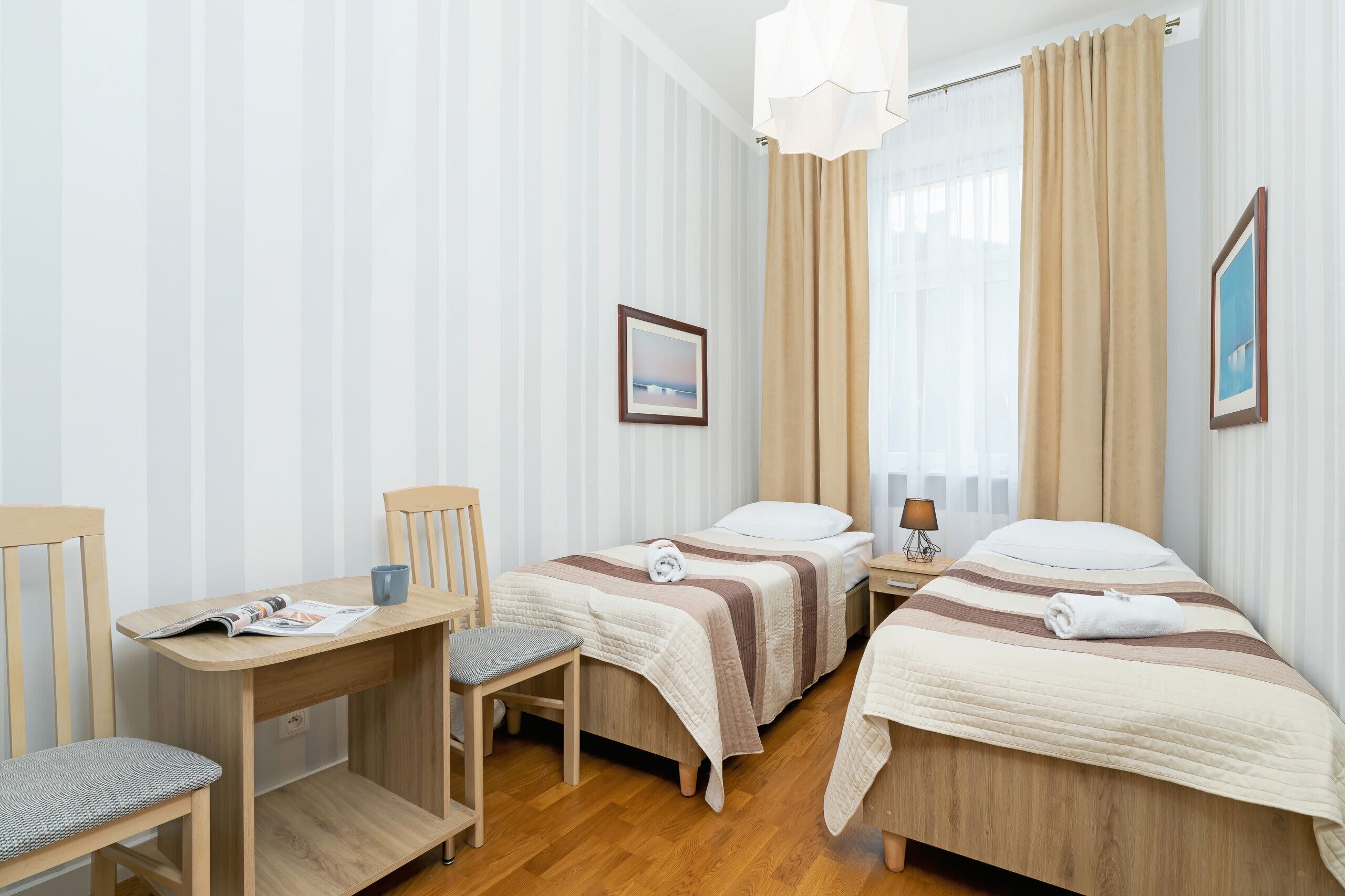 A hotel room with two single beds, a wooden desk with a chair, a small table with a lamp, and a picture on the wall.
