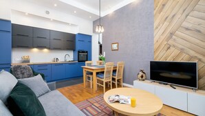 Una cocina moderna con armarios azules, un comedor con sillas de madera, un televisor sobre un mueble blanco y un sofá con un tapete estampado.
