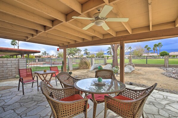 An outdoor patio with wicker chairs and a glass table, a ceiling fan, and a view of a landscaped area with palm trees and a fence. - Indio Escape w/ Fire Pit & Resort Amenities! (Indio)