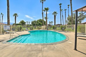 A swimming pool with a curved edge, surrounded by a fence, palm trees, and a clear blue sky. - Indio Escape w/ Fire Pit & Resort Amenities! (Indio)