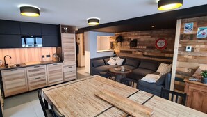 Une cuisine moderne avec des meubles noirs, des plans de travail en bois et un îlot central. On y trouve un coin repas avec une table et des chaises en bois. La pièce est agrémentée d’un mur d’accent en bois, décoré, et d’un petit coin salon avec un canapé et des coussins.