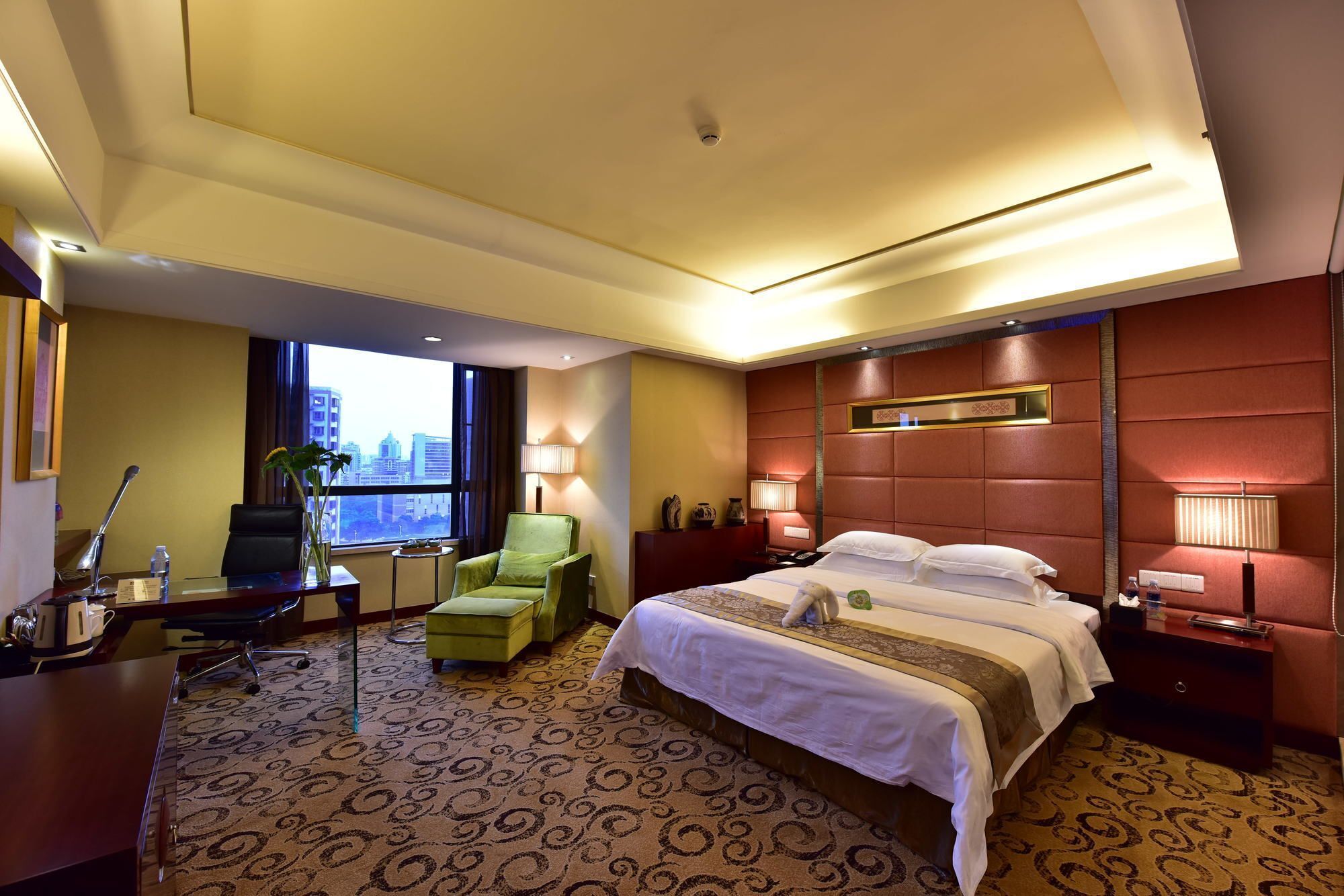 A hotel room with a large bed, a desk with a chair, a small table, a potted plant, and a view of the city through the window.