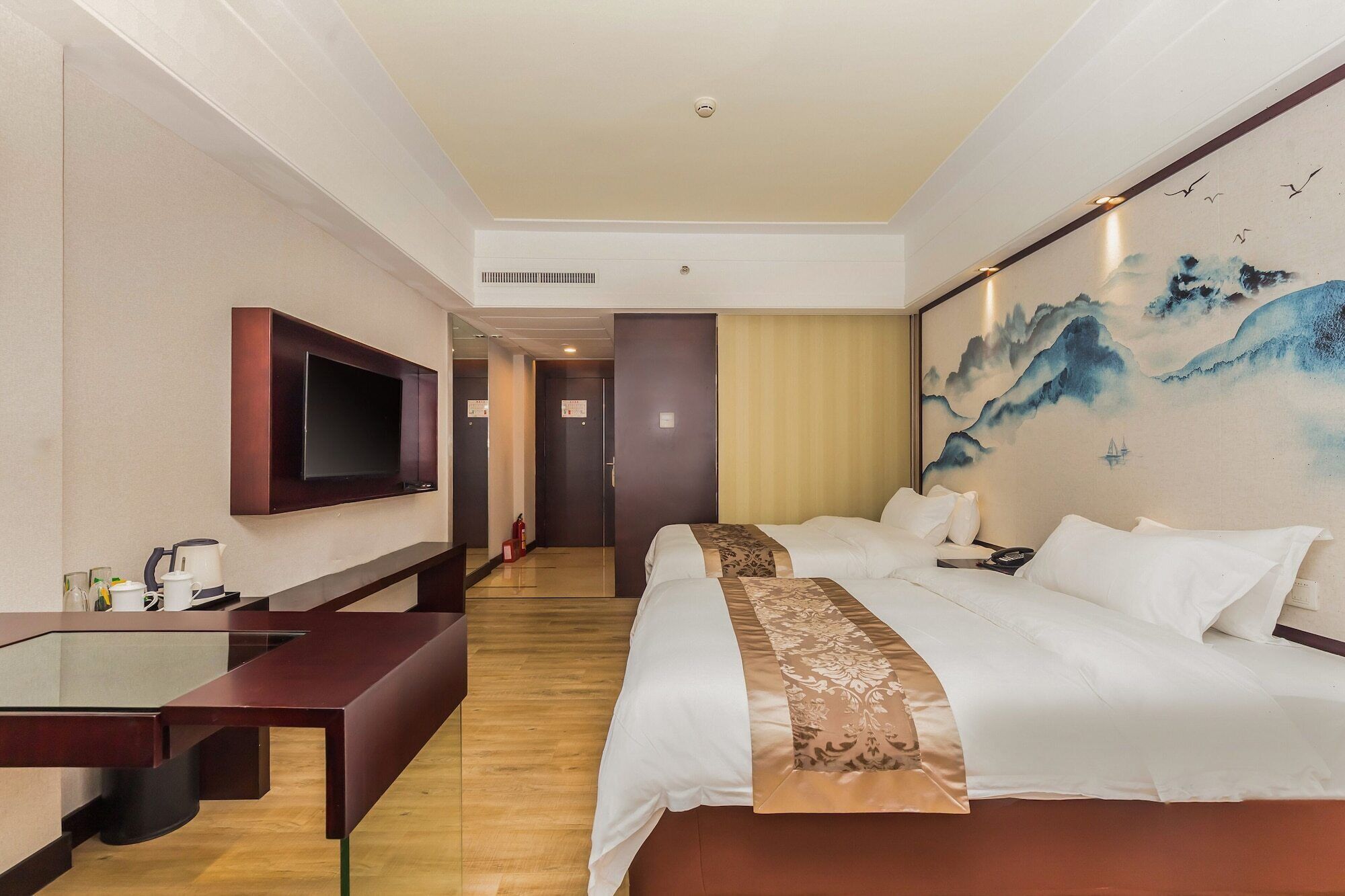 A hotel room with a large bed, a flat-screen TV mounted on the wall, a desk with a kettle, and a painting of mountains on the wall.