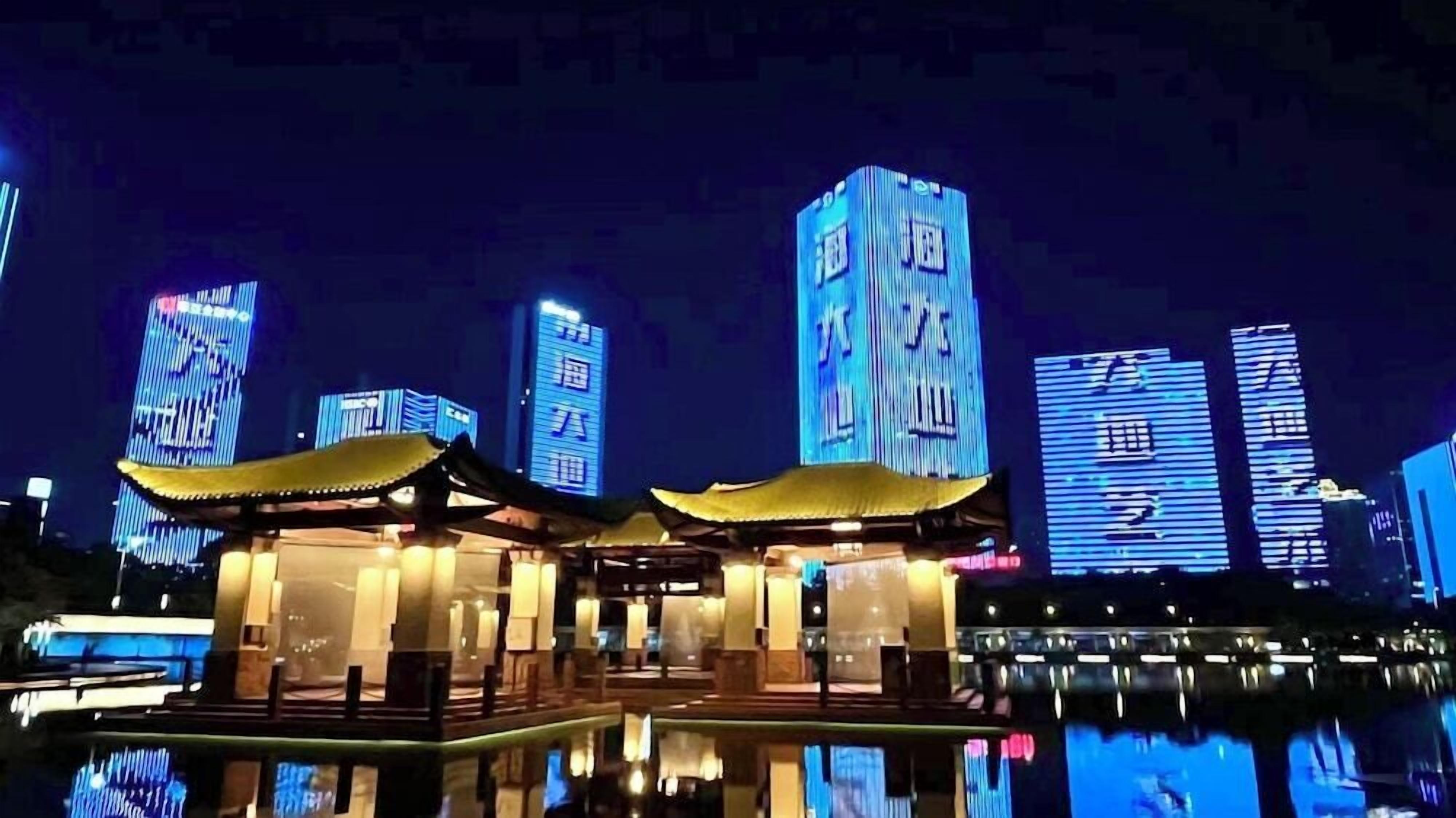A traditional pavilion with a reflective pool in front, surrounded by modern illuminated buildings at night.