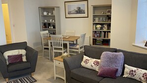 Ein Wohnzimmer mit grauem Sofa, einem Holztisch, einem Esstisch mit Stühlen und einem Bücherregal.