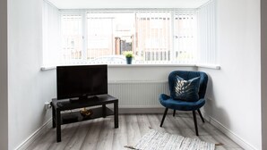A modern living room with a television on a black stand, a blue velvet chair, and a view of buildings through a large window. - Refurbished Belfast Home, 10 mins from centre with parking! (Belfast)
