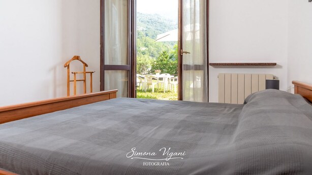 A bedroom with a large bed, a wooden chair, a radiator, and a view of greenery outside. - Feel at Home - LA TORRICELLA (LOVERE)