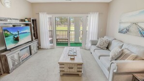 Een woonkamer met een televisie, een bank, een salontafel en uitzicht op een terras door glazen deuren.