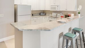 Een moderne keuken met witte kastjes, een marmeren aanrecht, een ingebouwde oven en een magnetron. Er staan twee barkrukken bij een klein eiland met een spoelbak en een wijnglas.