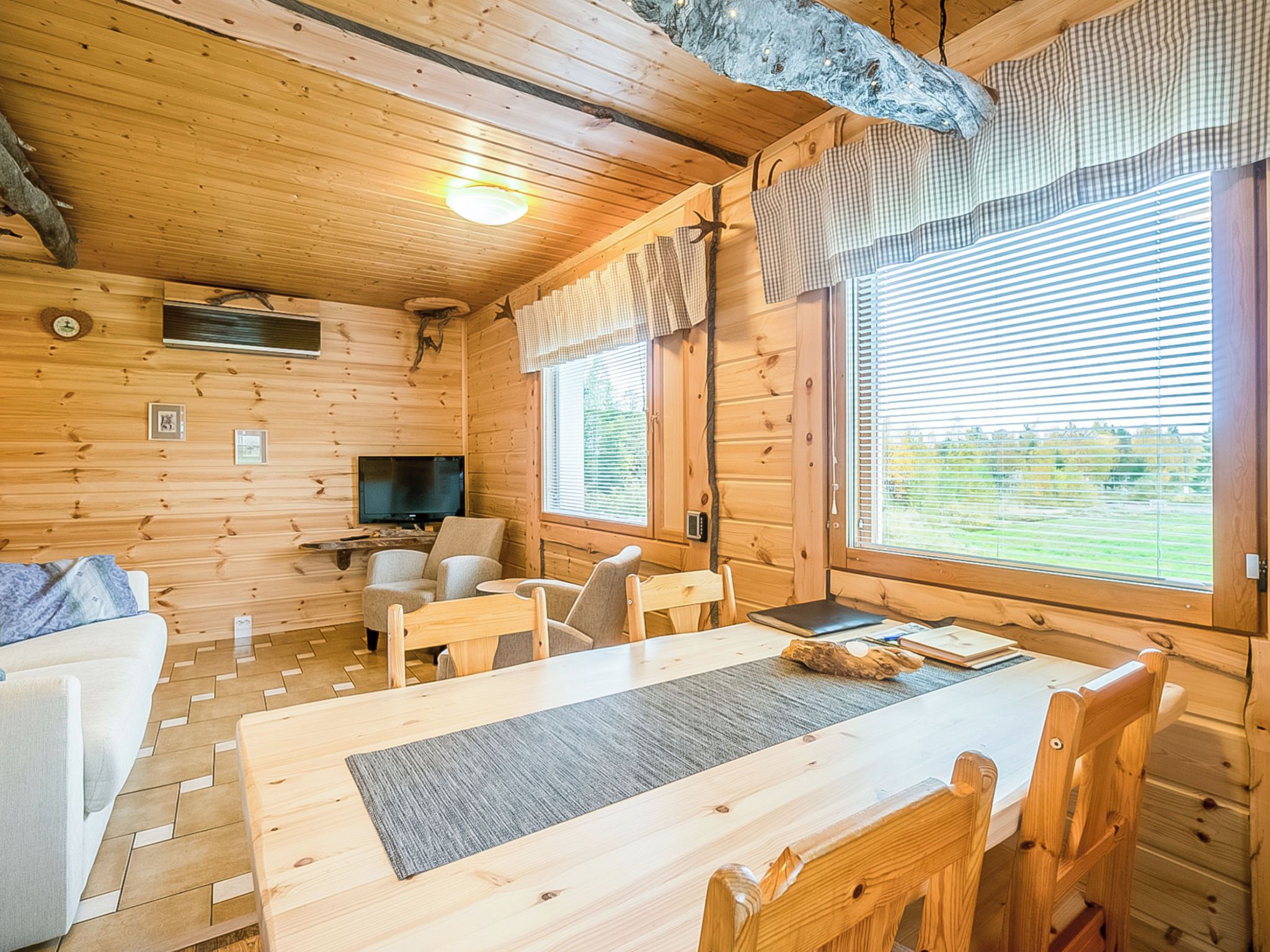 Ein Holzblockbau-Innenraum mit Esstisch, Stühlen, einer Couch, einem Fernseher und Fenstern mit Vorhängen.