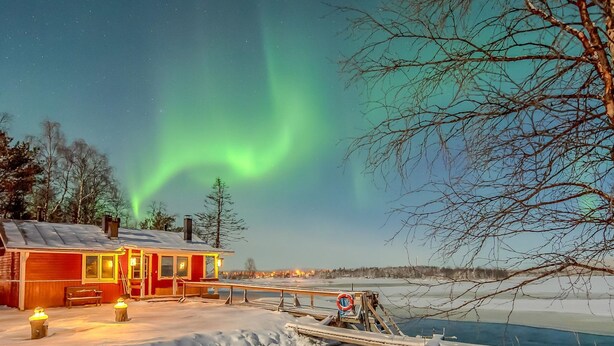 A red cabin with a dock extending into a frozen lake, surrounded by snow and trees.