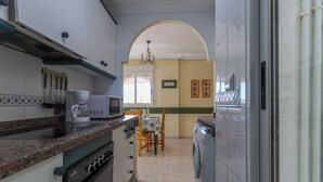 Una cocina equipada con microondas, cafetera y lavadora. Se observa una mesa de comedor con sillas y una lámpara de araña al fondo.