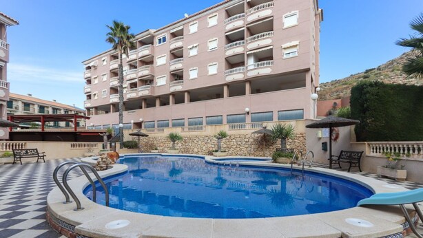 Una piscina con suelo a cuadros, rodeada por un muro de piedra y un edificio de varios pisos con balcones.