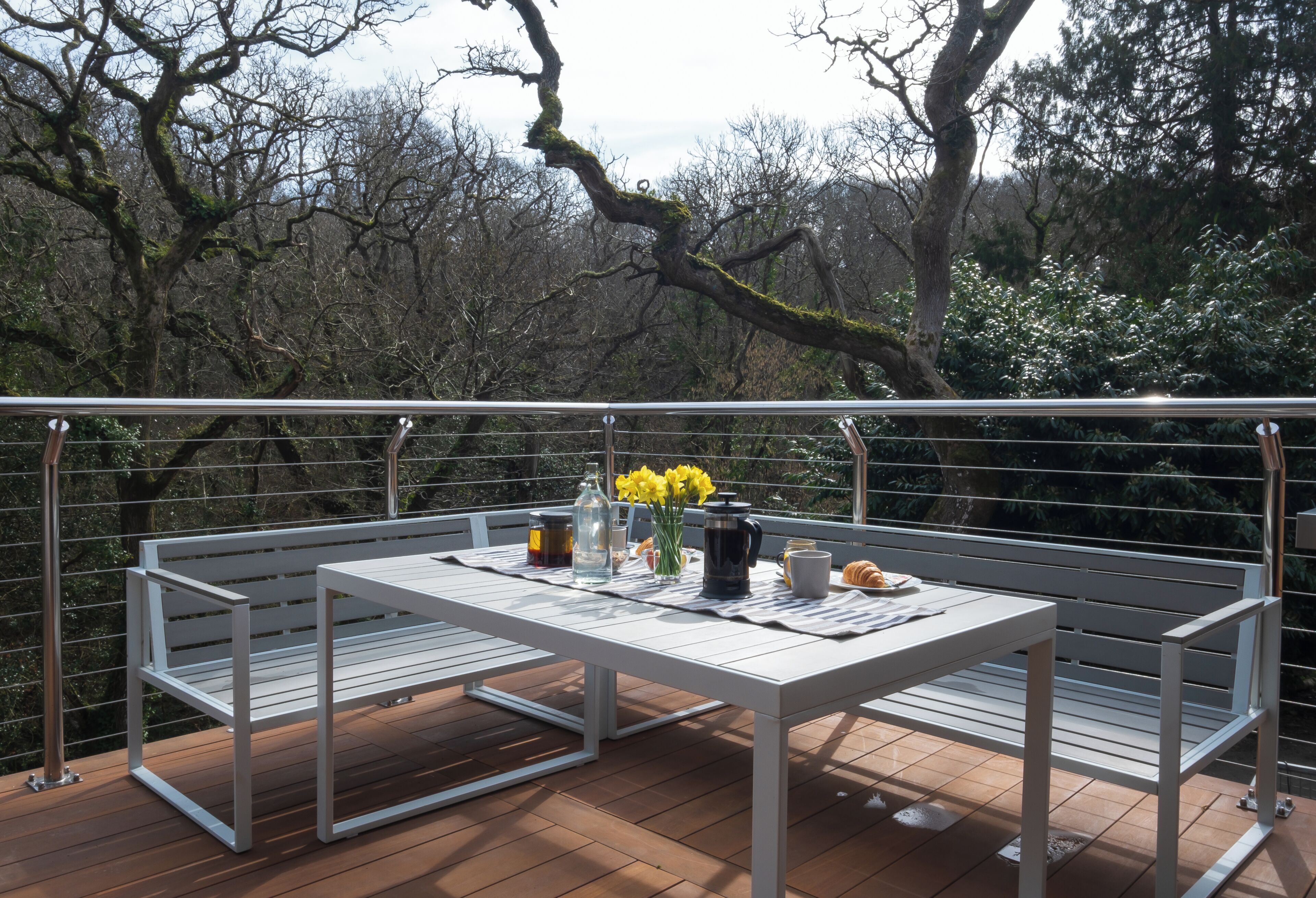 A balcony with a white table set for breakfast, including a coffee pot, mug, and flowers.