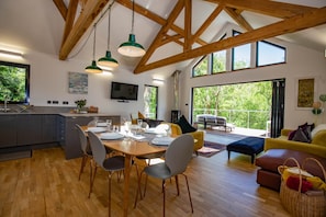 A spacious kitchen with a dining area, wooden beams, large windows, and a TV.