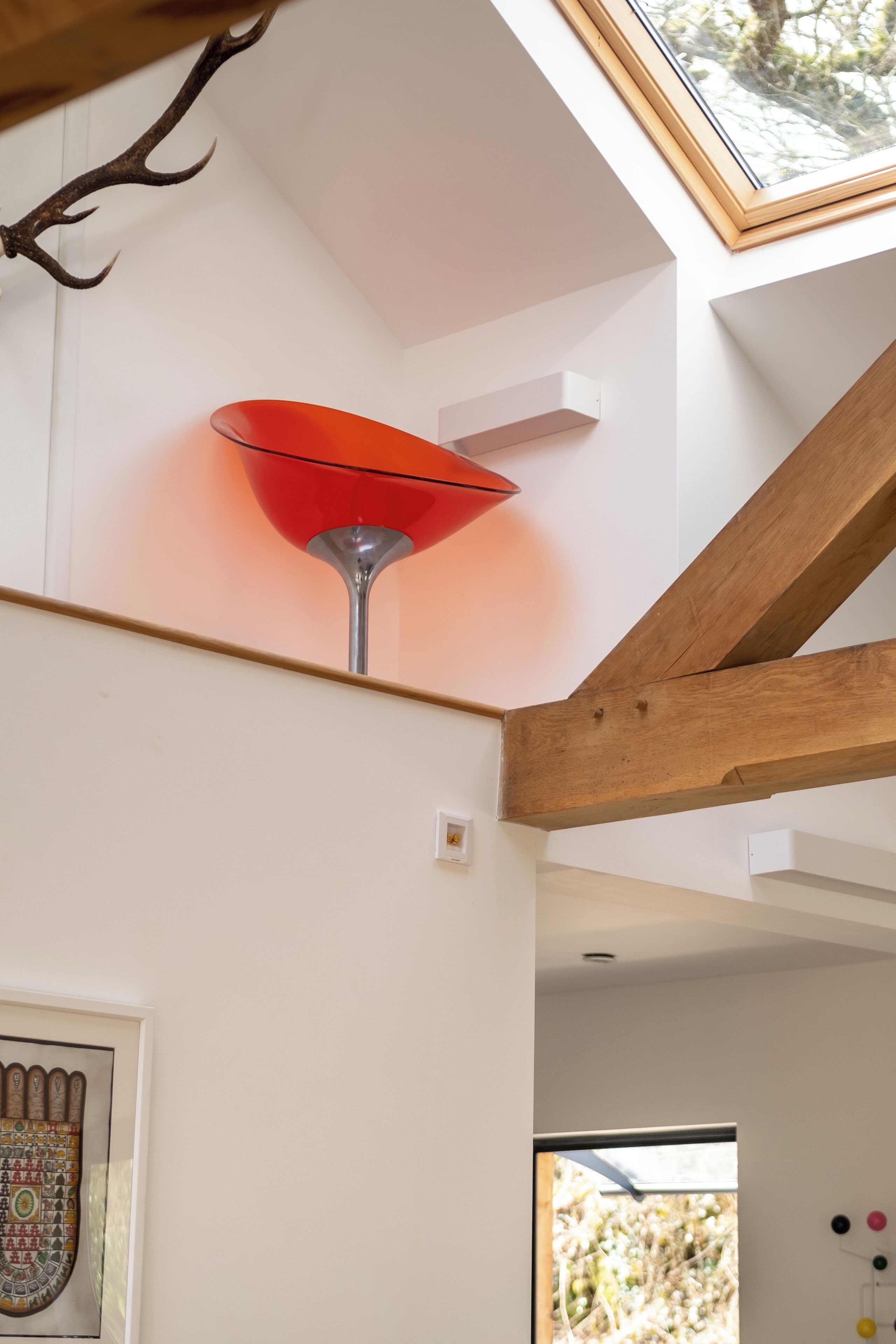 A room with a slanted ceiling, a red lamp, a framed artwork, and a window with a view of trees.