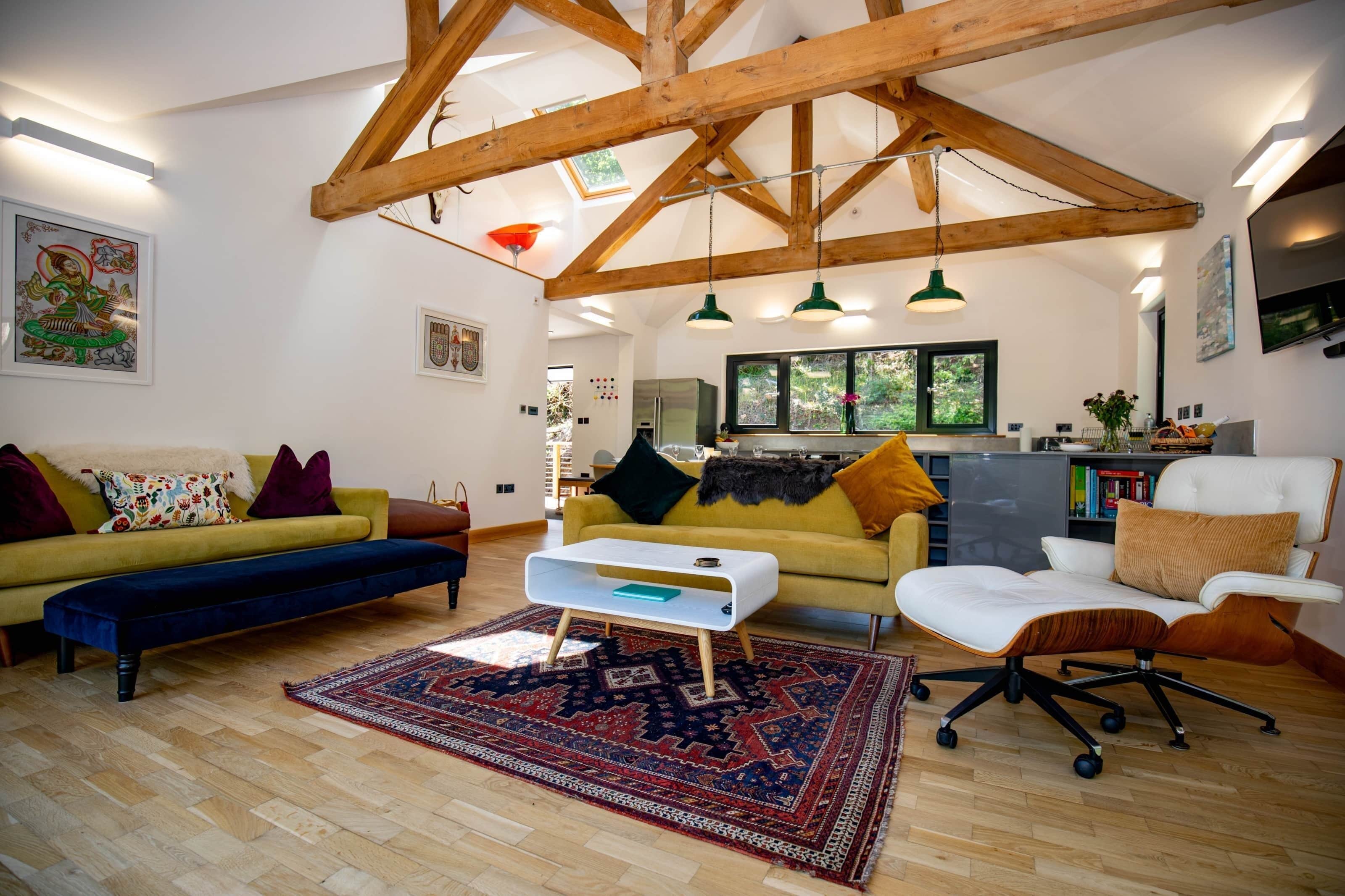 Ein geräumiges Wohnzimmer mit hoher Decke, Holzbalken, einer Sofagarnitur, einem Couchtisch und Blick zur Küche.