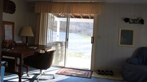 A room with a wooden desk, a chair, a lamp, a large window with curtains, a door leading outside, and a small rug on the floor.