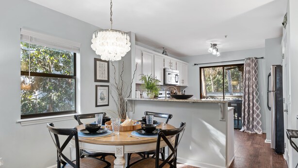 A dining area with a round table, chairs, and a chandelier. There is a kitchen area with cabinets and appliances in the background.