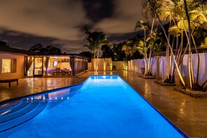 Une piscine dont le bord est illuminé de lumière bleue, entourée de palmiers et dotée d’un espace extérieur couvert avec une table et des chaises.