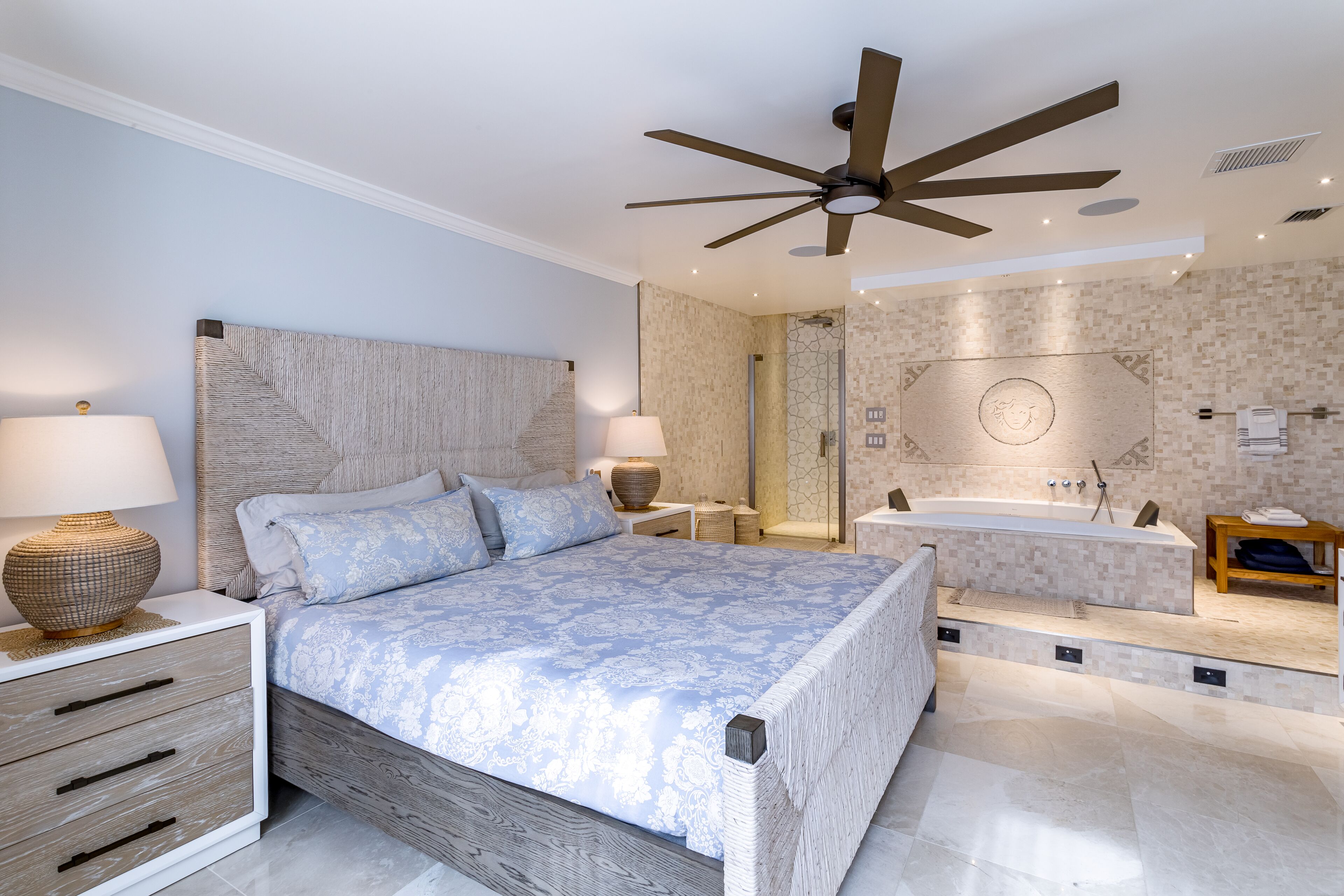 Une chambre moderne dotée d’un grand lit, de lampes de chevet, d’un ventilateur de plafond, d’un dressing et d’une salle de bain avec baignoire et douche.