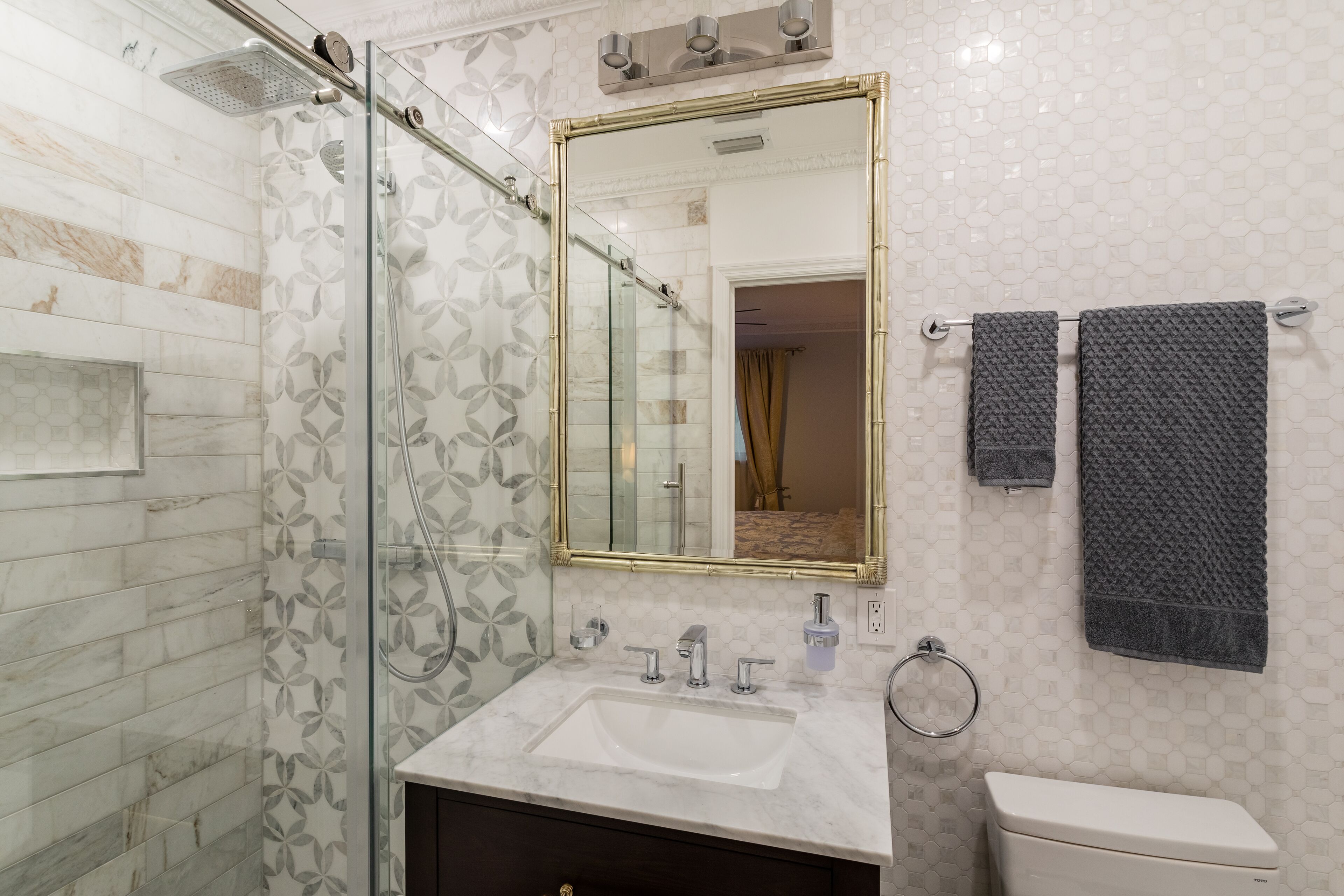 A bathroom with a glass shower enclosure, a white sink, a toilet, and a mirror with a gold frame.