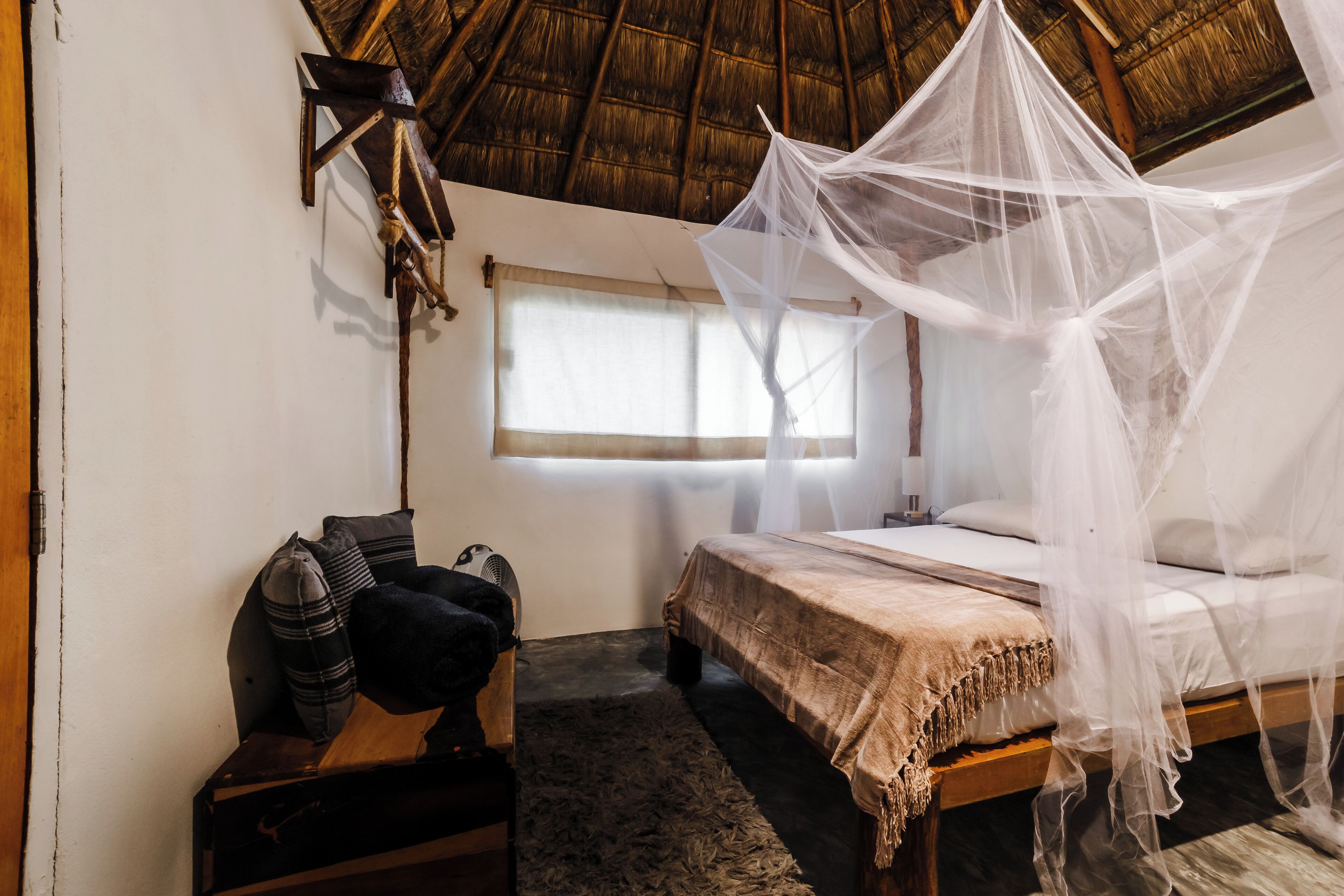 A room with a bed under a mosquito net, a wooden headboard, a window with curtains, and a fan on a wooden table.