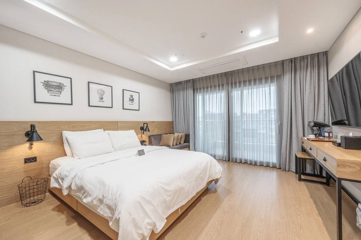 A hotel room with a bed, bedside lamps, a desk, and a large window with curtains.