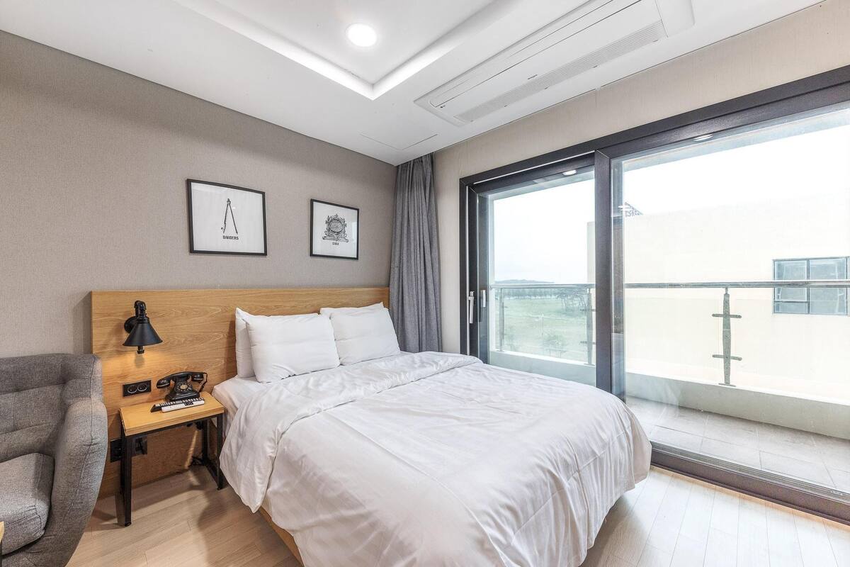 A hotel room with a bed, a desk with a lamp, a chair, and a large window with a view.