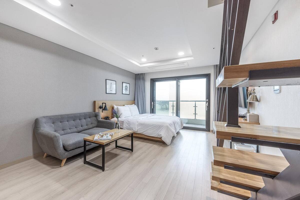 A modern hotel room with a grey sofa, a wooden coffee table, a bed with white bedding, and a staircase leading to an upper floor.