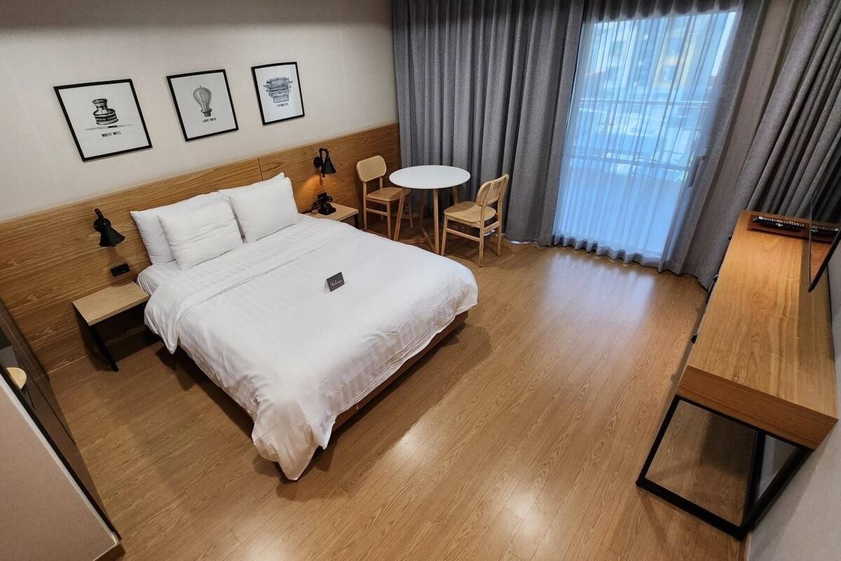 A hotel room with a large bed, bedside tables, a small round table, two chairs, a desk with a chair, and a window with curtains.