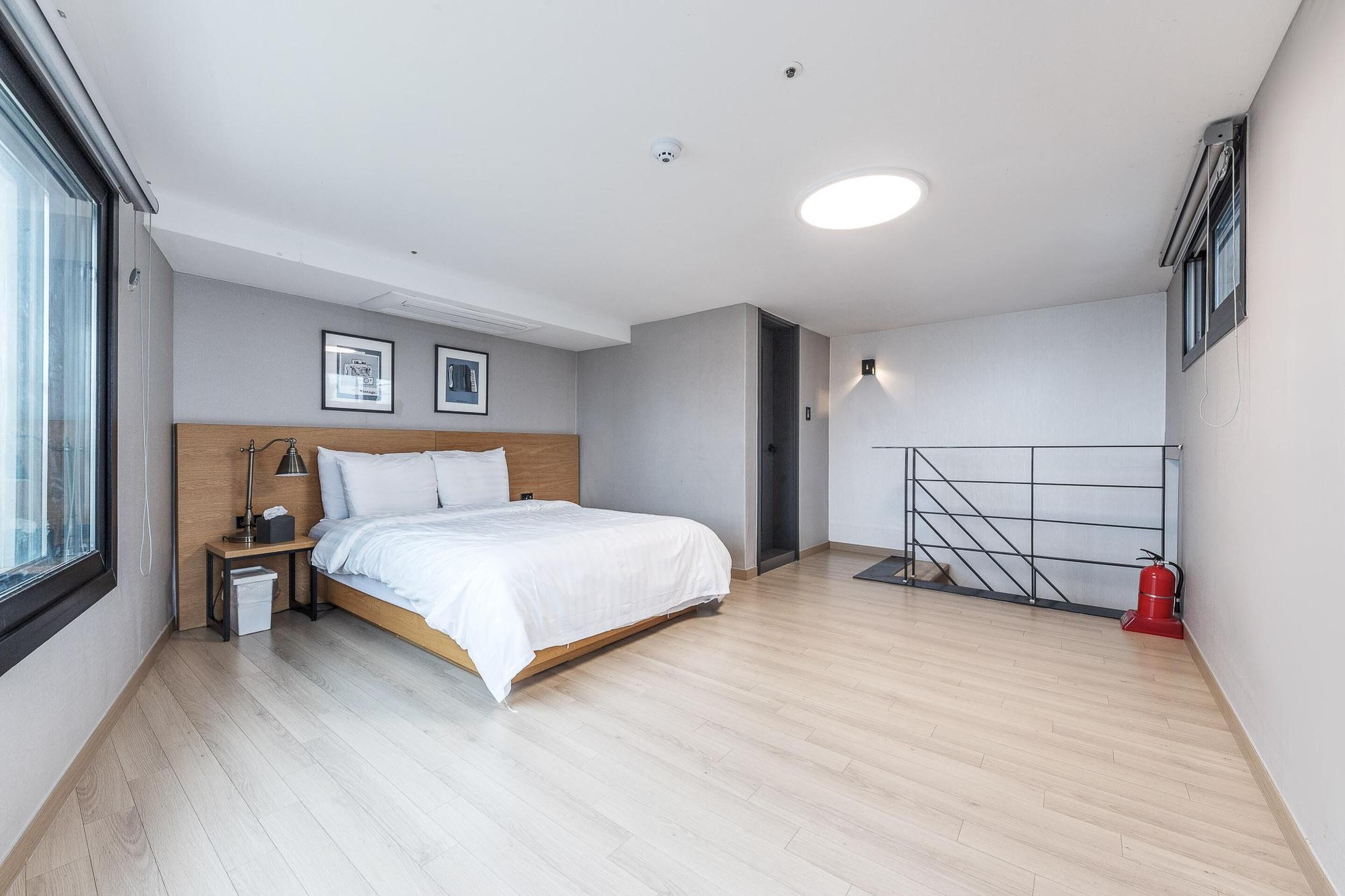 Een moderne hotelkamer met een houten bed, nachtkastje, bedlampje, een kleine prullenbak, een brandblusser en een raam met uitzicht.