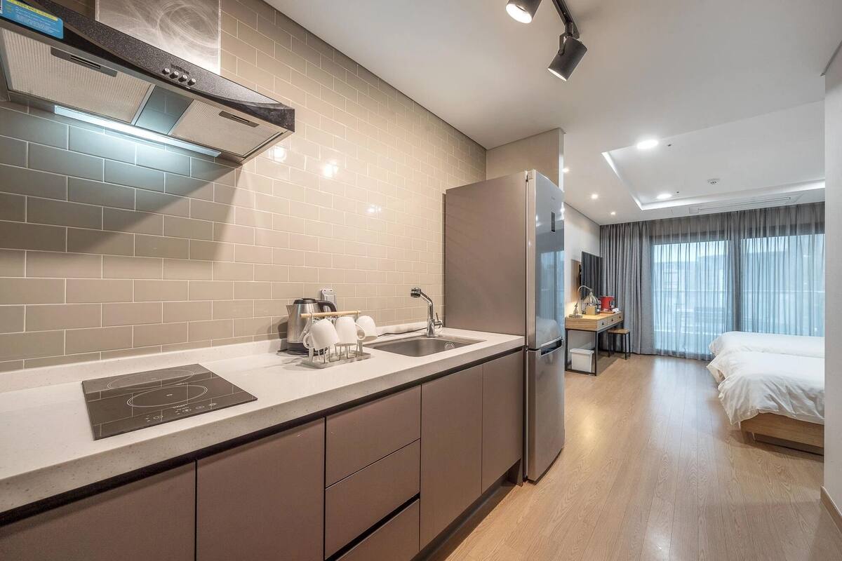 A modern hotel room with a kitchenette, a bed, a desk, and a large window.