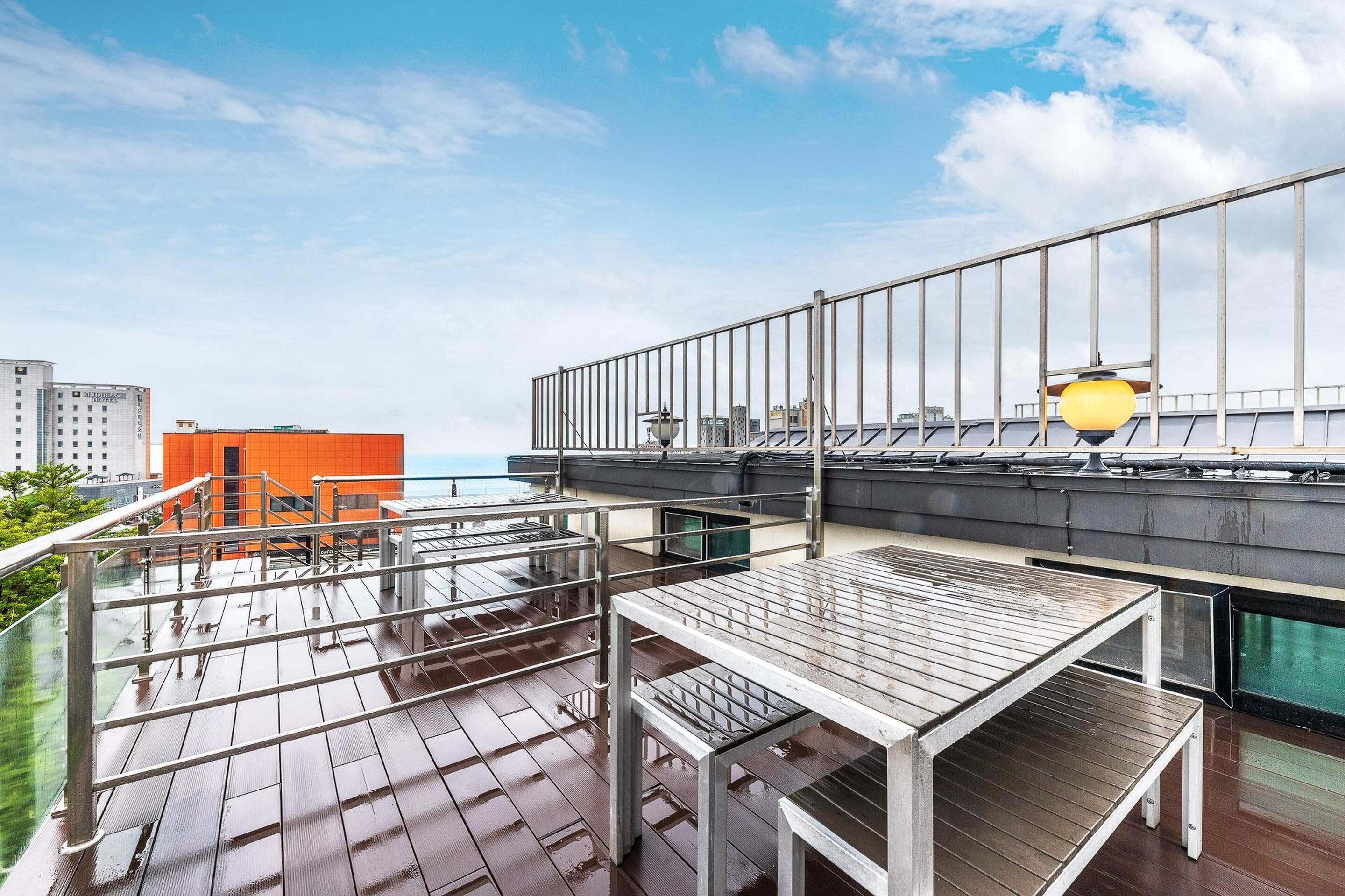 A rooftop terrace with wooden tables and chairs, a metal railing, and a view of the cityscape and ocean.