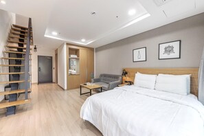 A modern hotel room with a wooden staircase, a bed with white linens, a gray armchair, a small table, and a wooden cabinet. - Daecheon 2278 Resort (Boryeong)
